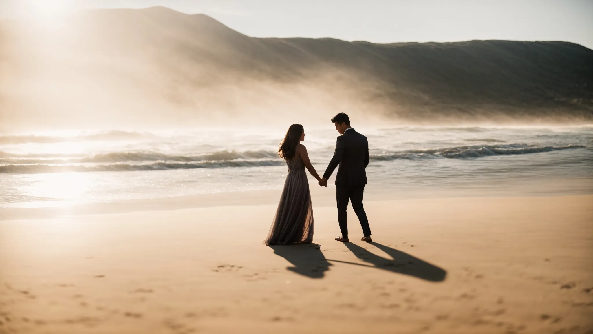 a couple embraces on a sunlit beach, a silhouetted photographer captures their moment from a short distance.