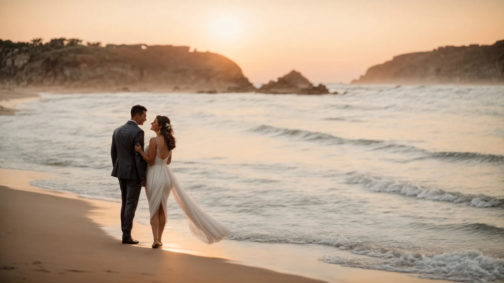 a bride and groom walk on a beach at sunset, pausing to share a kiss as the photographer captures the moment.