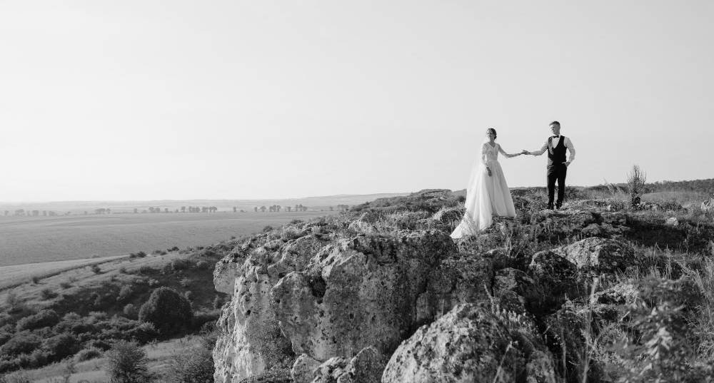 Black-and-white photograph of a bride and groom holding hands on a rocky hillside