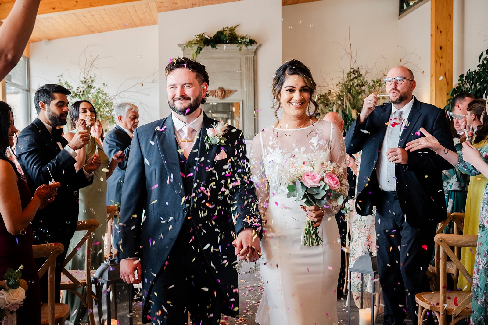 Bride and groom walking through colourful confetti inside a timber-framed venue