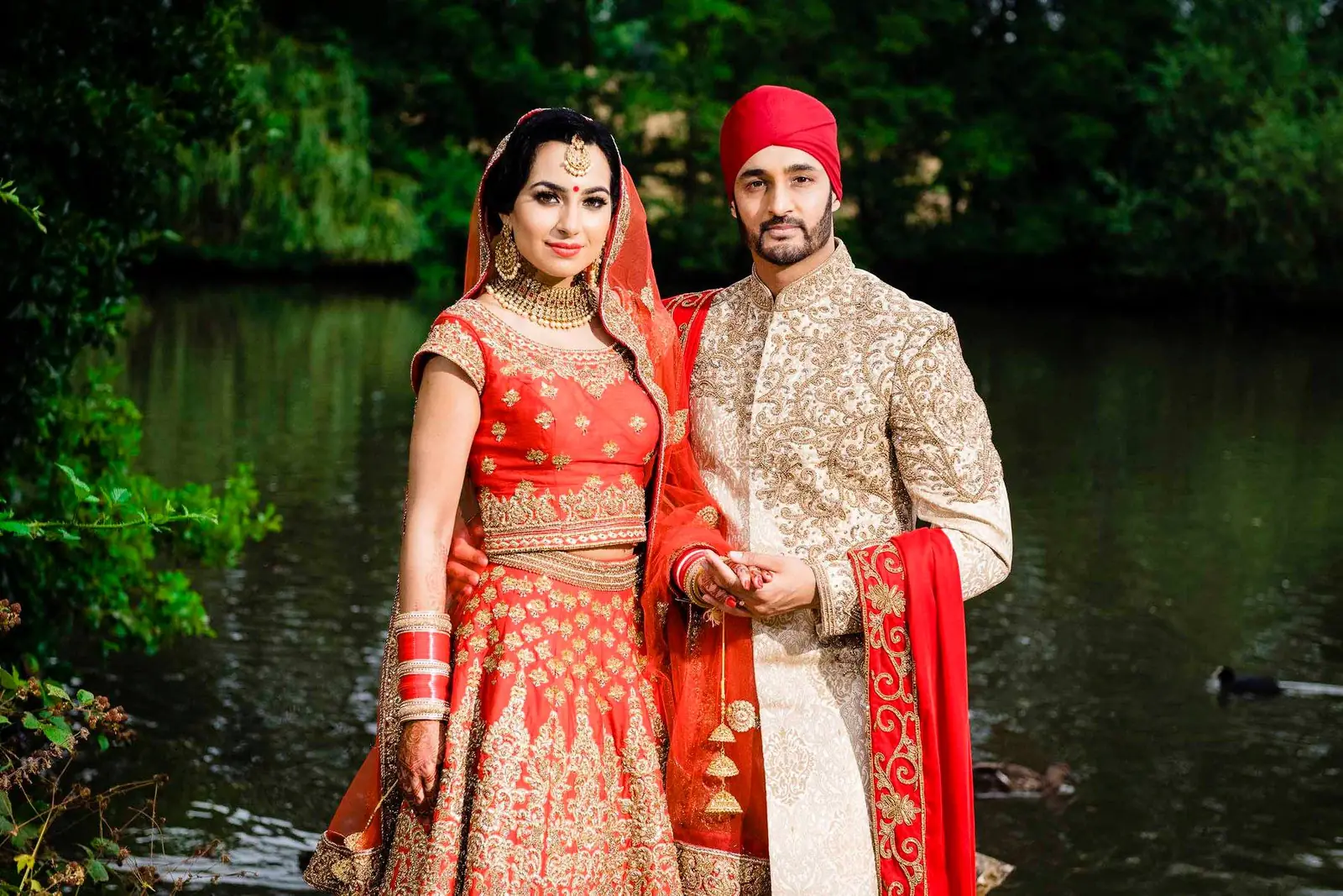 Couple in red and gold wedding attire beside a pond