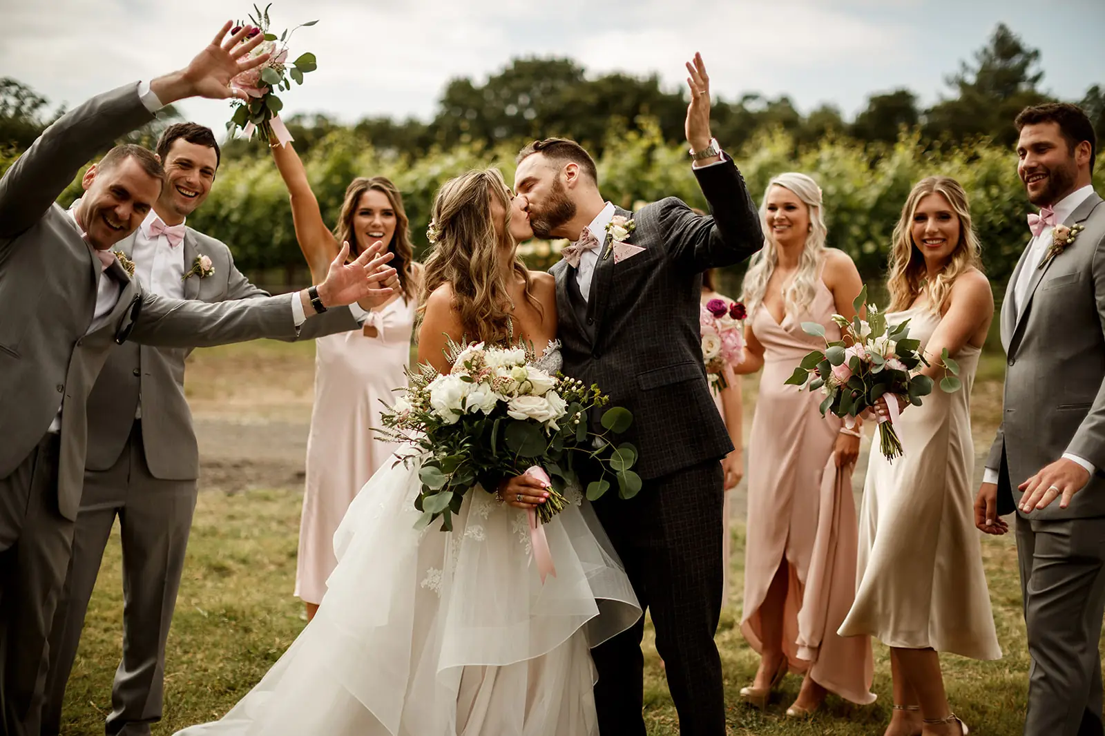 Newlyweds kissing outdoors while their wedding party celebrates