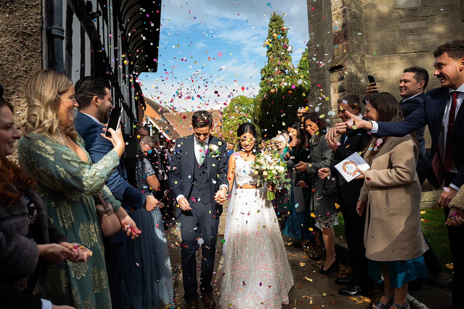Destination wedding confetti exit Newlyweds holding hands as guests shower them with colourful confetti outside a stone church