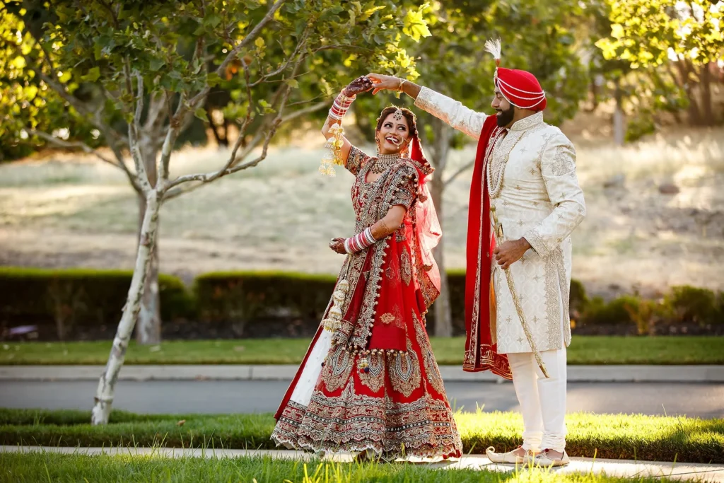 Bride in a red embroidered lehenga twirling beneath the groom’s raised hand in a sunlit garden