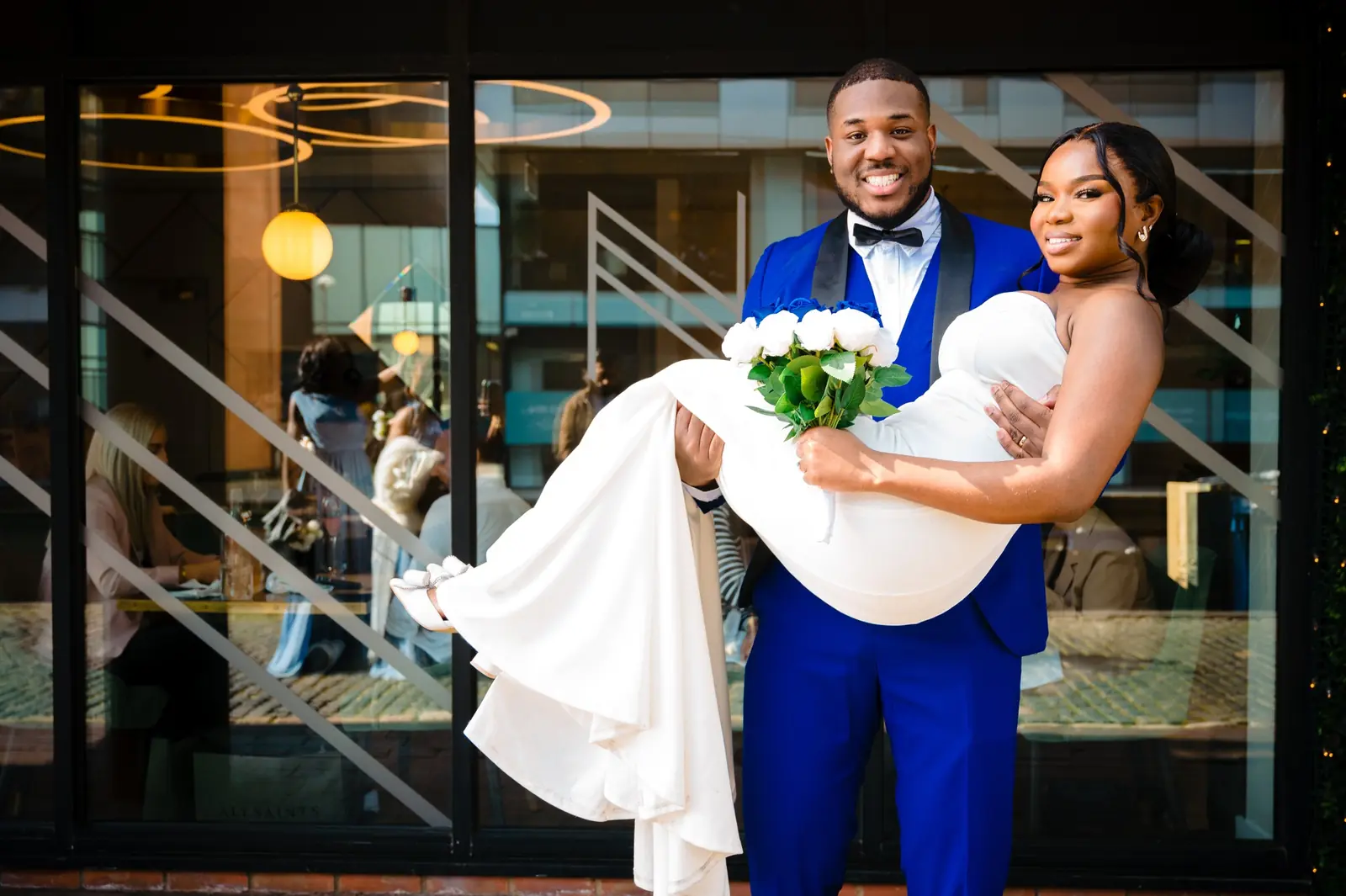 Groom in a blue suit carrying his smiling bride outside their wedding venue