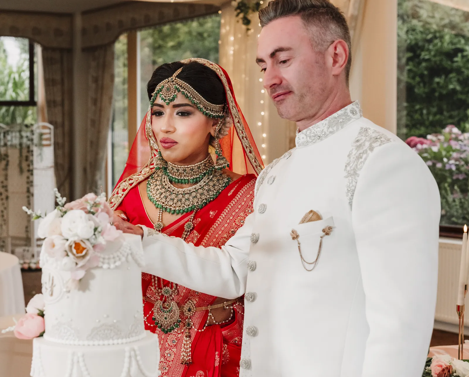 Bride in a red saree and groom in a white sherwani cutting their wedding cake at a fusion wedding